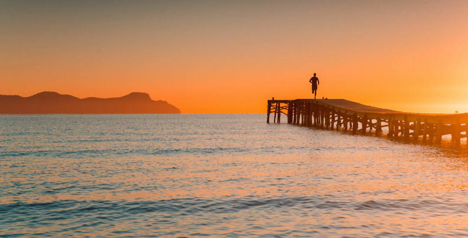 A person stands at the end of a wooden pier overlooking a calm sea during sunset. The sky is painted in warm orange and yellow hues, and distant, silhouetted mountains create a serene backdrop. The water gently ripples towards the shore.