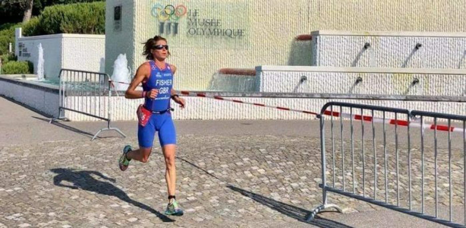An athlete in a blue triathlon suit and sunglasses runs past a barricade on a paved path. Behind them is a white wall with fountains and the words "CIO/IOC" and "Le Musée Olympique" inscribed on it. The weather appears sunny.