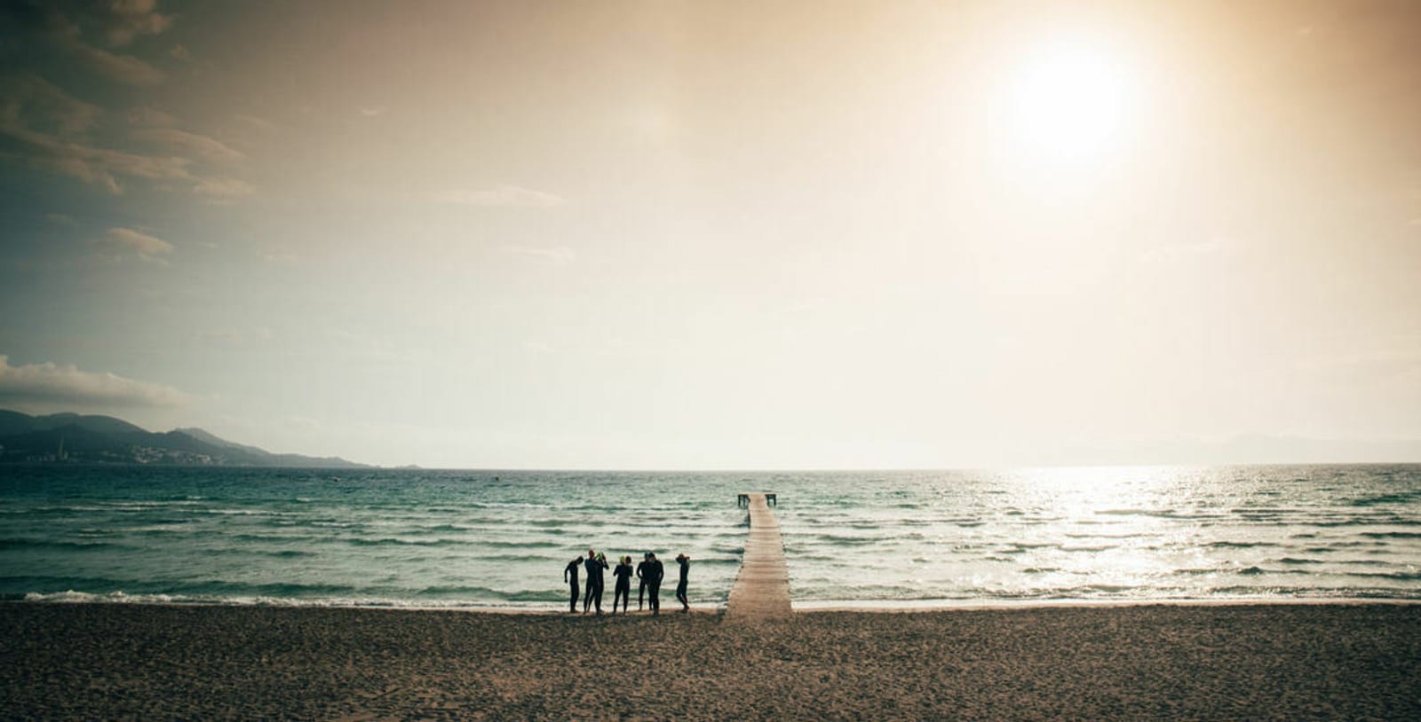 A group of people stands on a sandy beach near the edge of a wooden pier that extends into a calm sea. The sun is setting or rising, casting a warm glow over the water and sky, creating a serene and picturesque scene. Hills are faintly visible in the background.