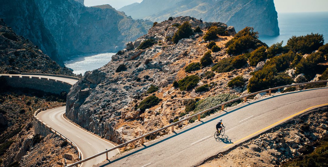 A cyclist rides along a winding road through a rocky coastal landscape with high cliffs and a distant view of the sea and mountainous terrain in the background under a clear sky.