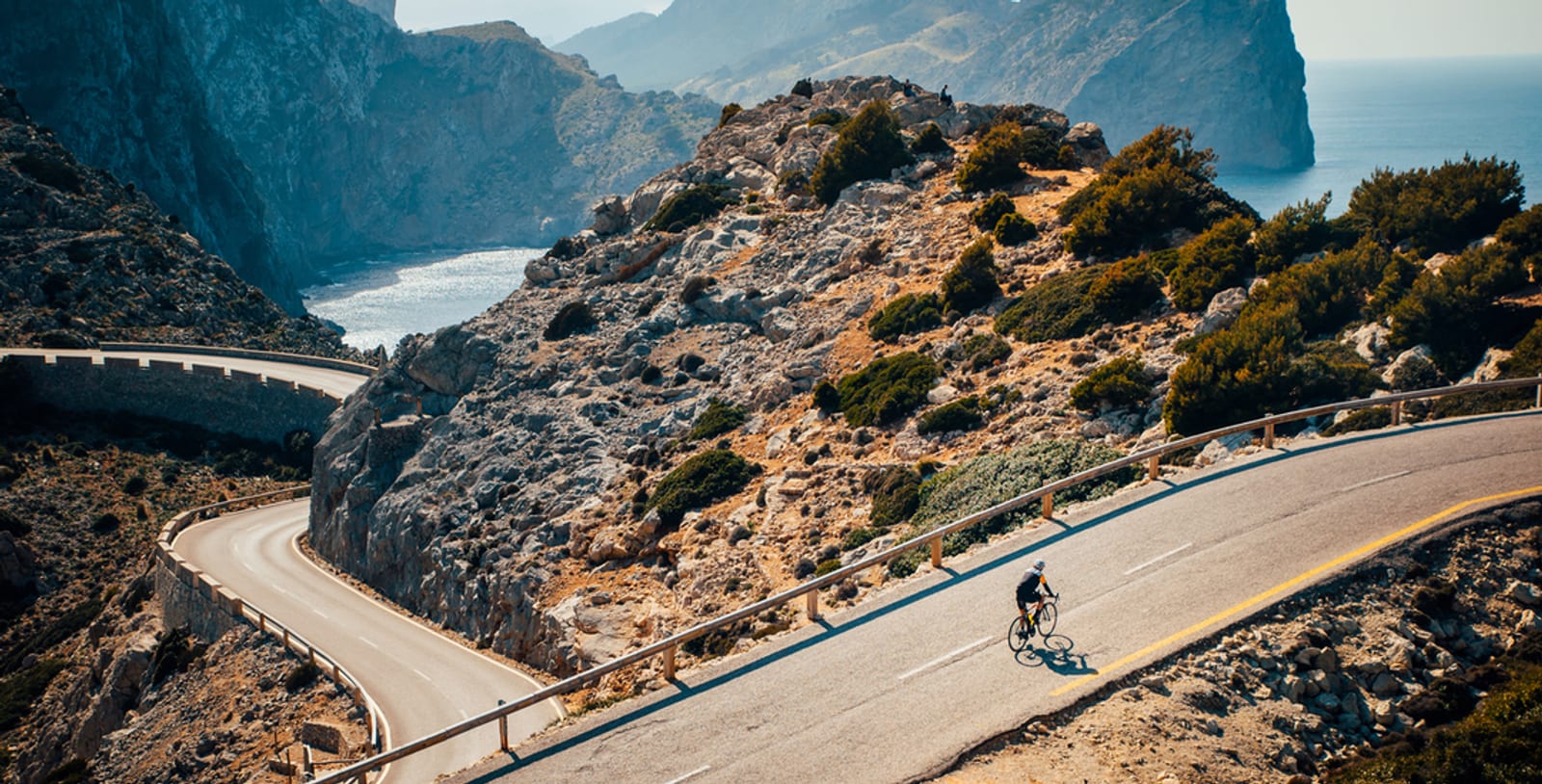 A cyclist rides along a winding road through a rocky coastal landscape with high cliffs and a distant view of the sea and mountainous terrain in the background under a clear sky.