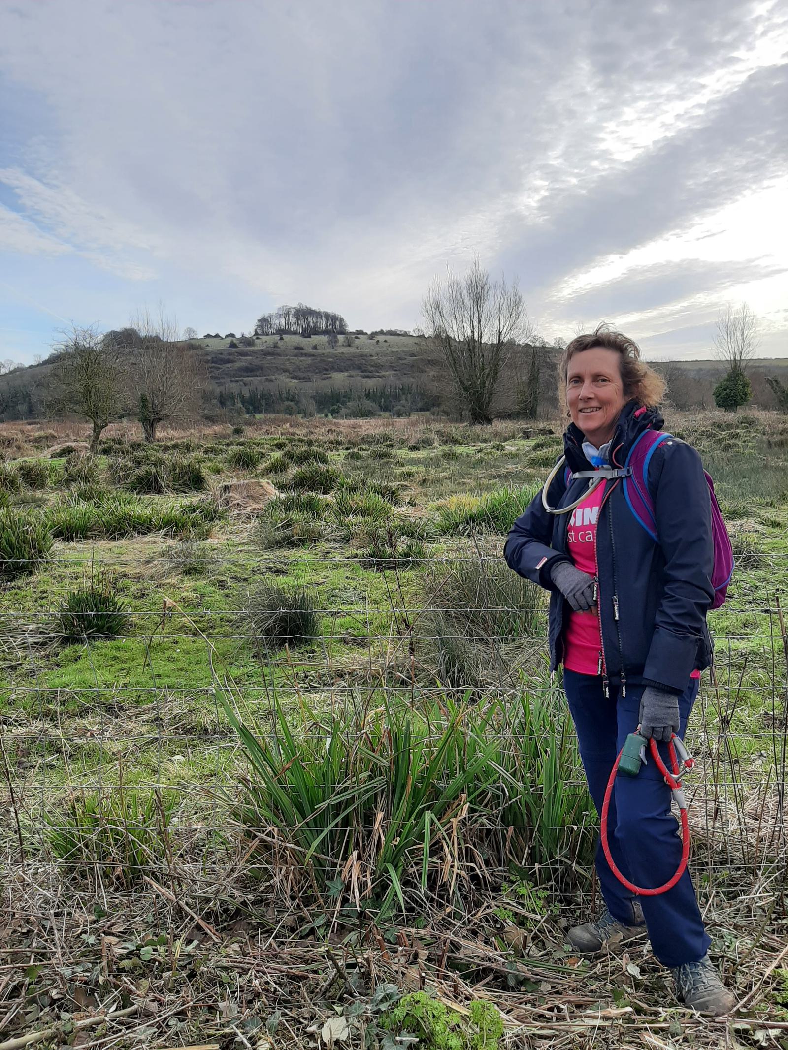 A person stands in a green field holding a red dog leash, with a small hill in the background. They are dressed in outdoor clothing, including gloves, and carry a purple backpack. The sky above is partly cloudy.