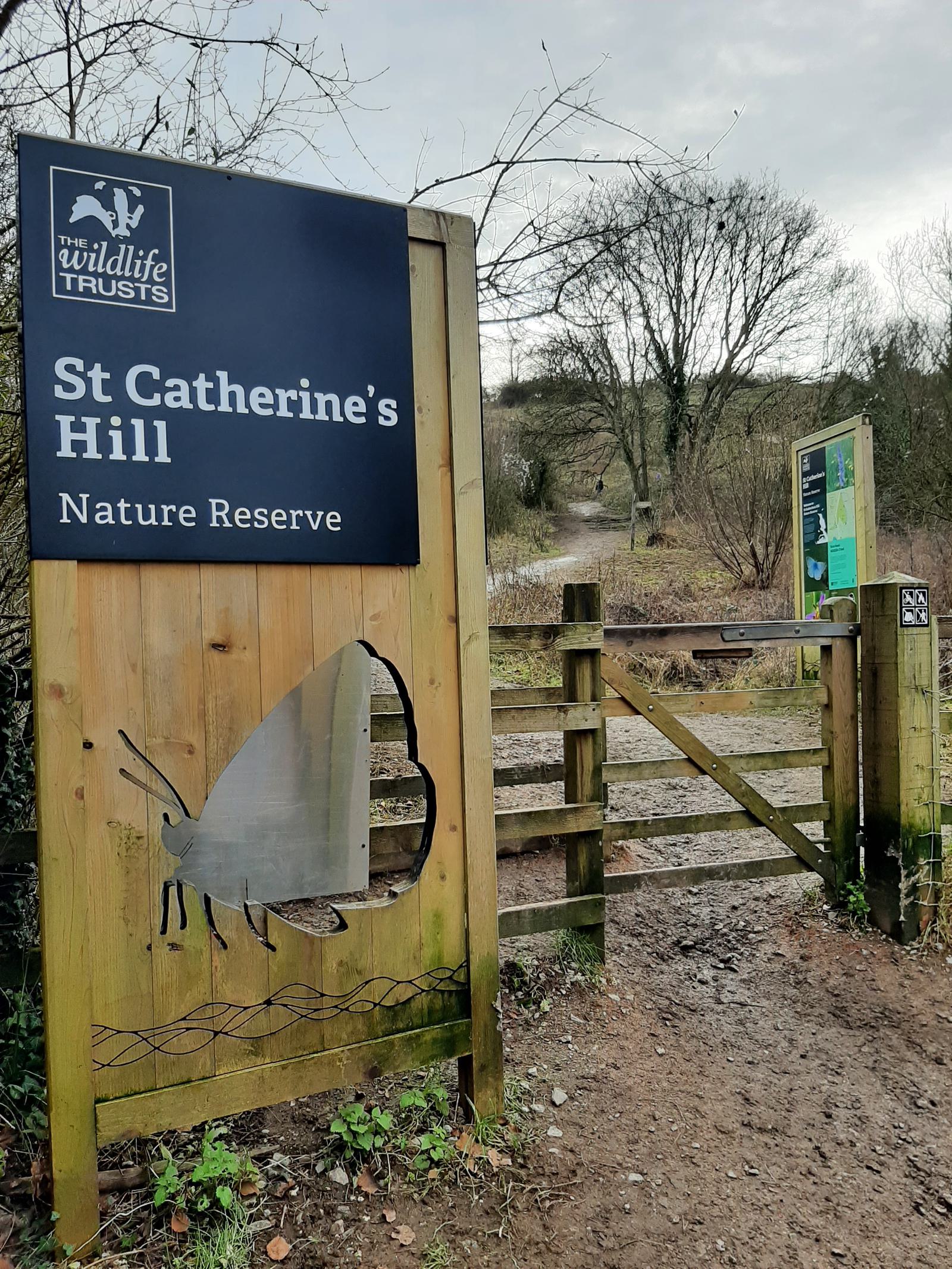 Entrance to St Catherine's Hill Nature Reserve, featuring a wooden gate and informational signs. The main sign shows a logo for The Wildlife Trusts and a butterfly silhouette. A dirt path leads into a wooded area with trees and overcast skies in the background.