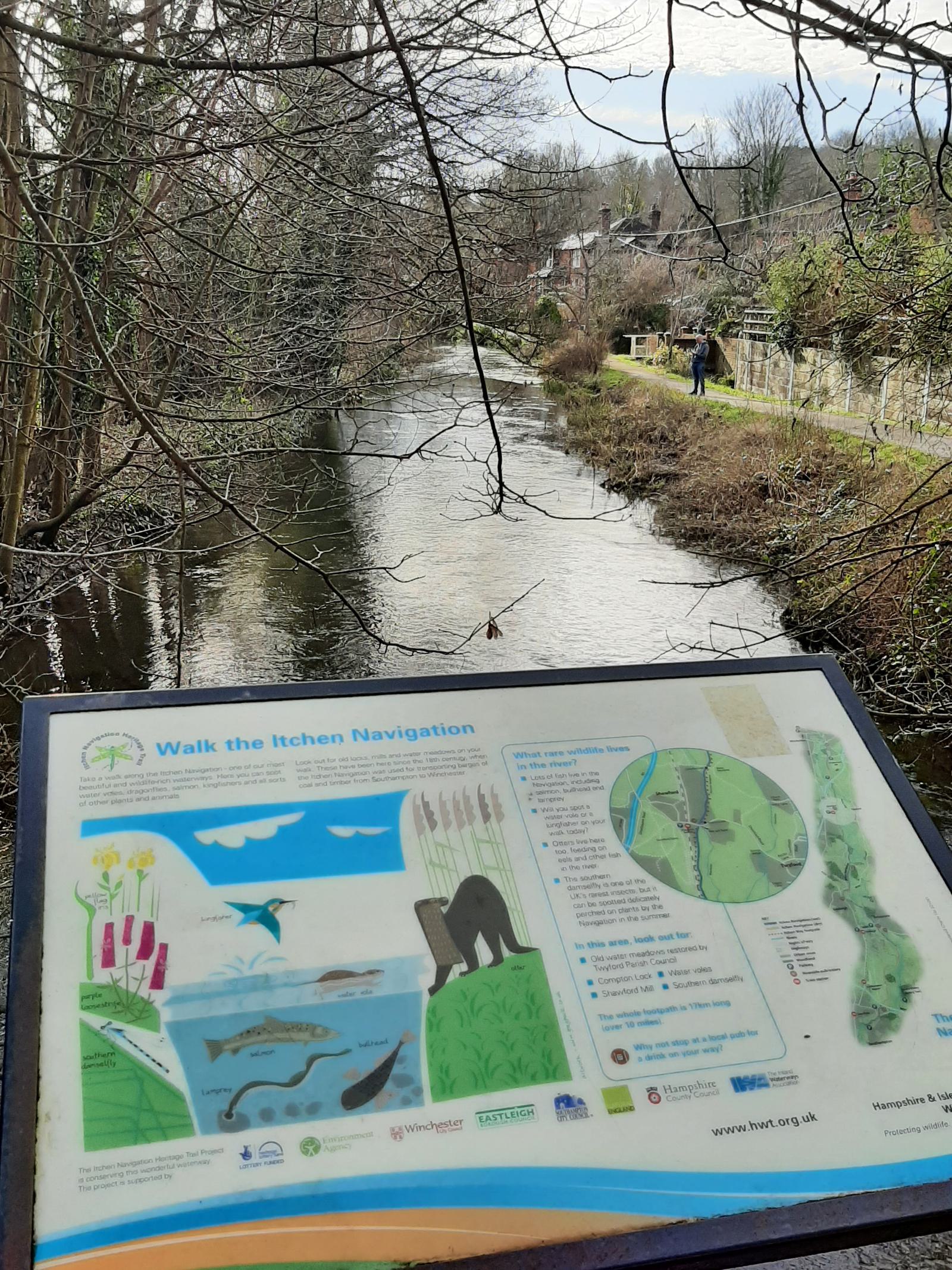 An informational sign about the "Walk the Itchen Navigation" trail stands beside a calm, narrow river lined with trees and shrubs. The background includes a walking path with a few people, houses, and overcast skies.
