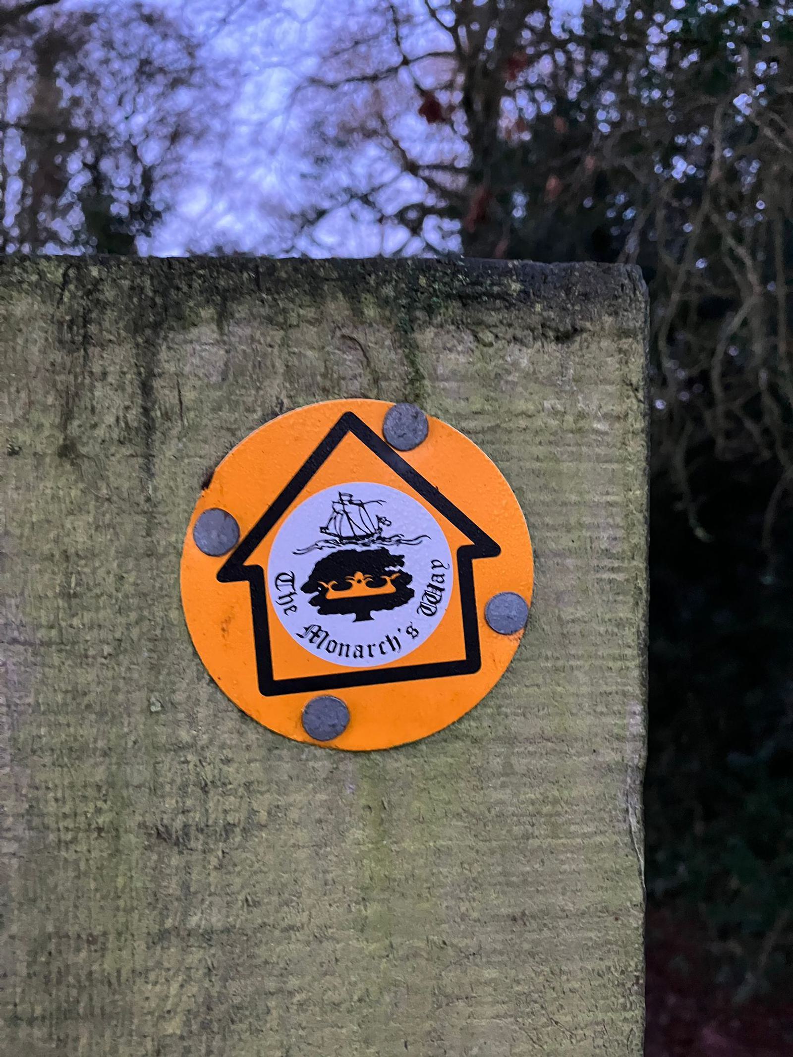A yellow circular trail marker with a black arrow on a wooden post. The arrow contains an image of a ship sailing above a crown with the text "The Monarch's Way." The background includes blurred foliage and a darkening sky.