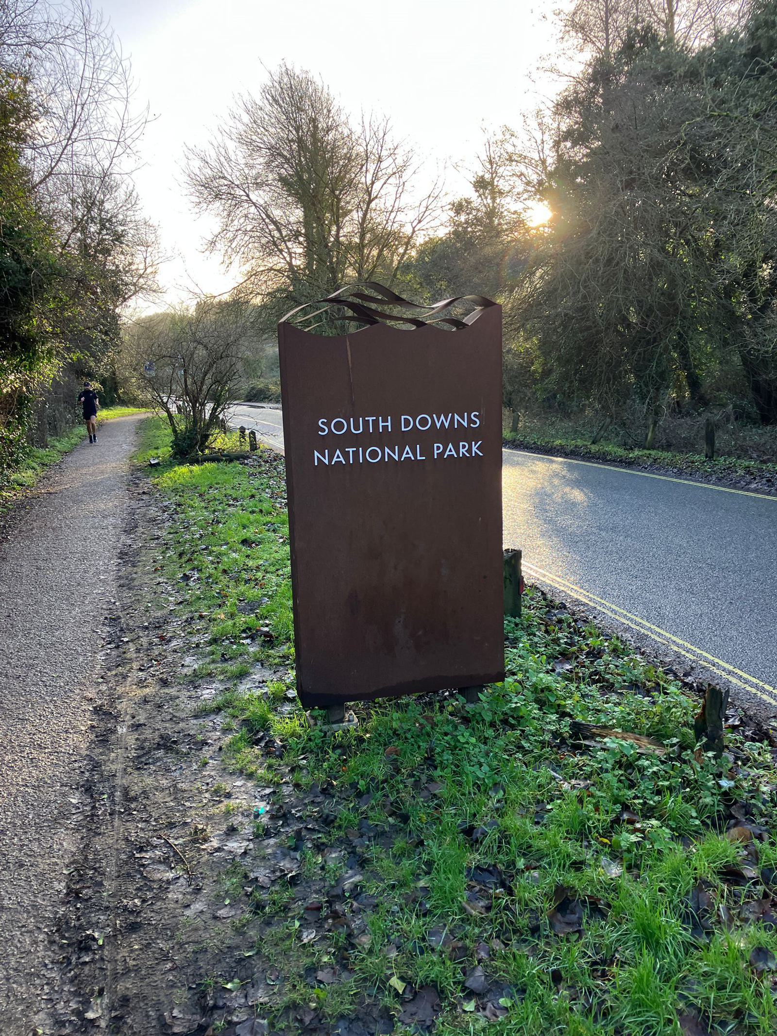 A sign for South Downs National Park stands beside a paved road and a dirt path in a wooded area. The sign is made of metal with white text. Sunlight filters through the trees, creating a serene atmosphere. A person is seen walking on the path in the distance.