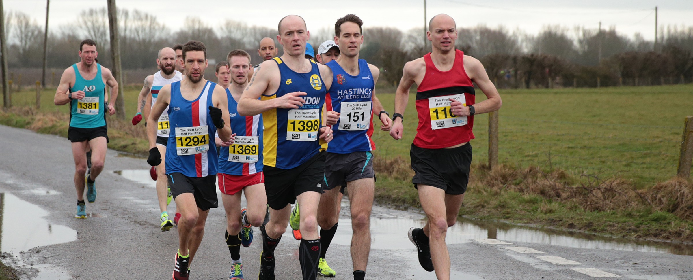 A group of male runners competing in a road race. They are wearing numbered bibs and a mix of running gear. The scene is set on a rural road with visible puddles and a green, overcast landscape. The runners look focused as they move forward.