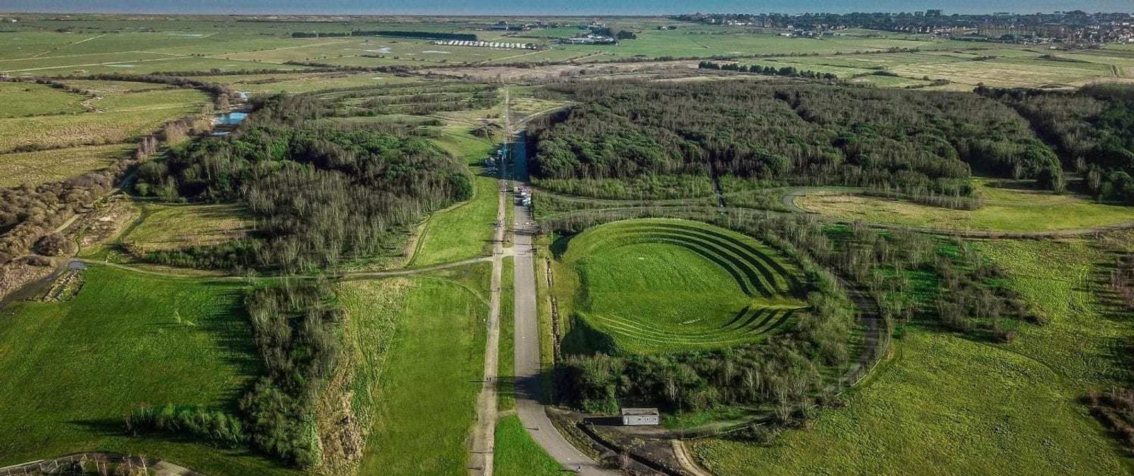 Aerial view of a park with a mix of open grasslands, dense tree areas, and a large, sculpted, grassy amphitheater-like structure. Paths and roads intersect the park, with surrounding fields and distant buildings on the horizon.