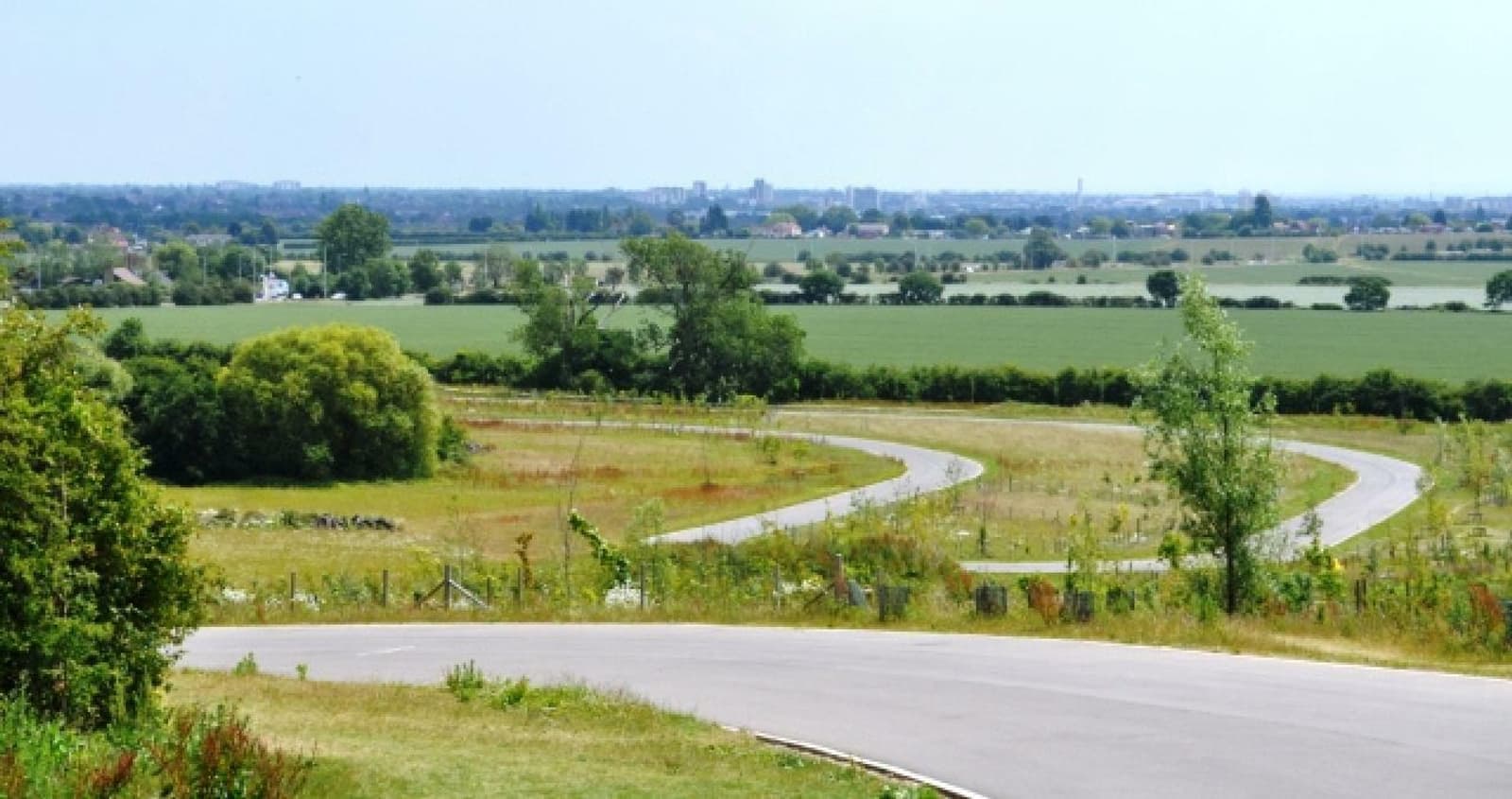 A scenic view of a winding road cutting through a lush, green countryside. The road is surrounded by fields and trees, with a distant cityscape visible on the horizon under a clear blue sky.