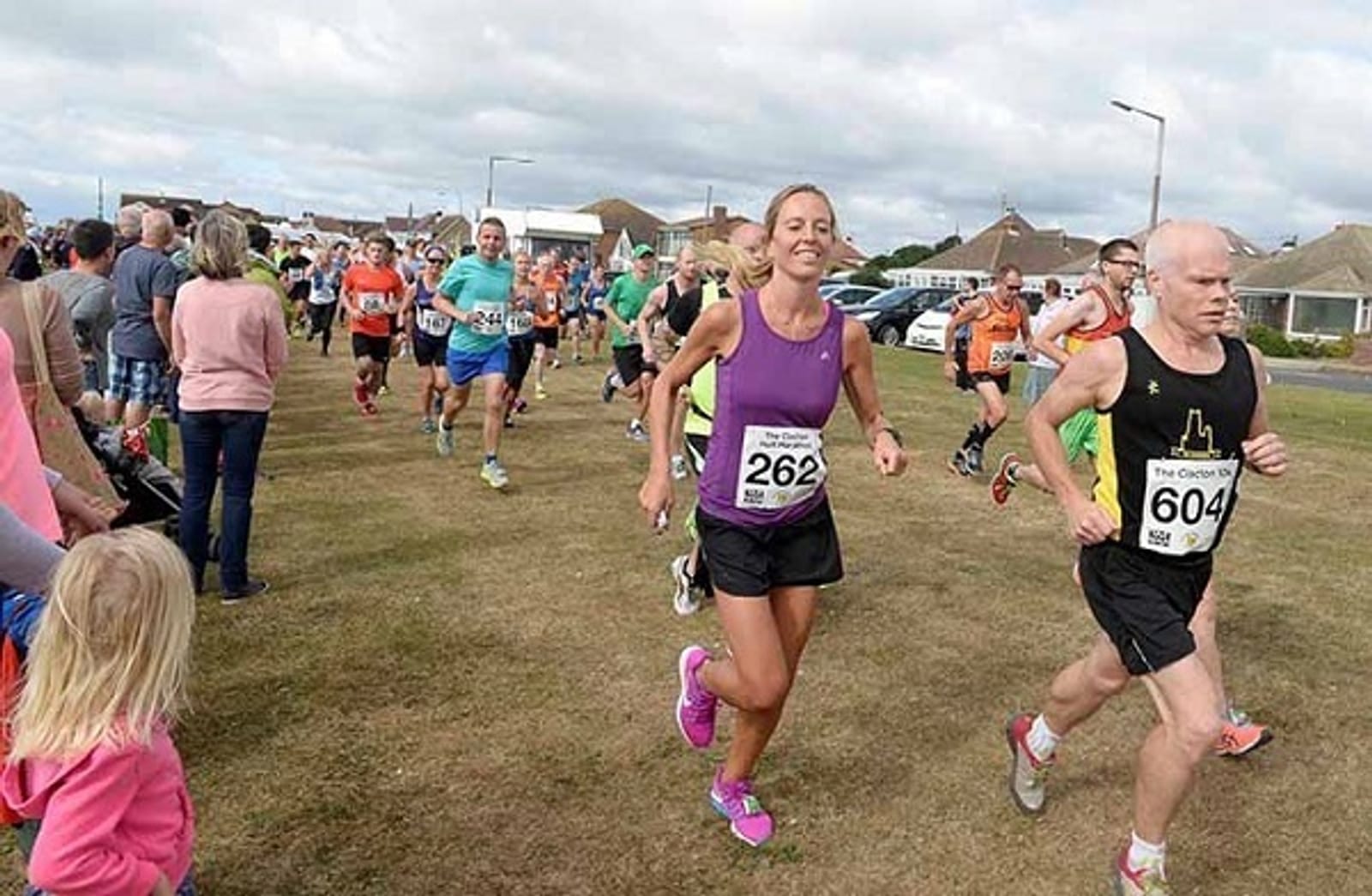 A group of runners participating in a race on a grassy field. One runner, wearing a purple tank top and black shorts, is numbered 262, while another, in a black tank top and shorts, is numbered 604. Spectators watch from the side, and houses are visible in the background.