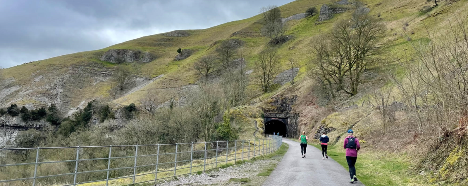 A group of people walking or running along a paved path that leads up to a tunnel in a hillside. The surrounding landscape features grassy slopes, scattered bare trees, and rocky outcrops under a cloudy sky. A metal fence lines the path on the left side.