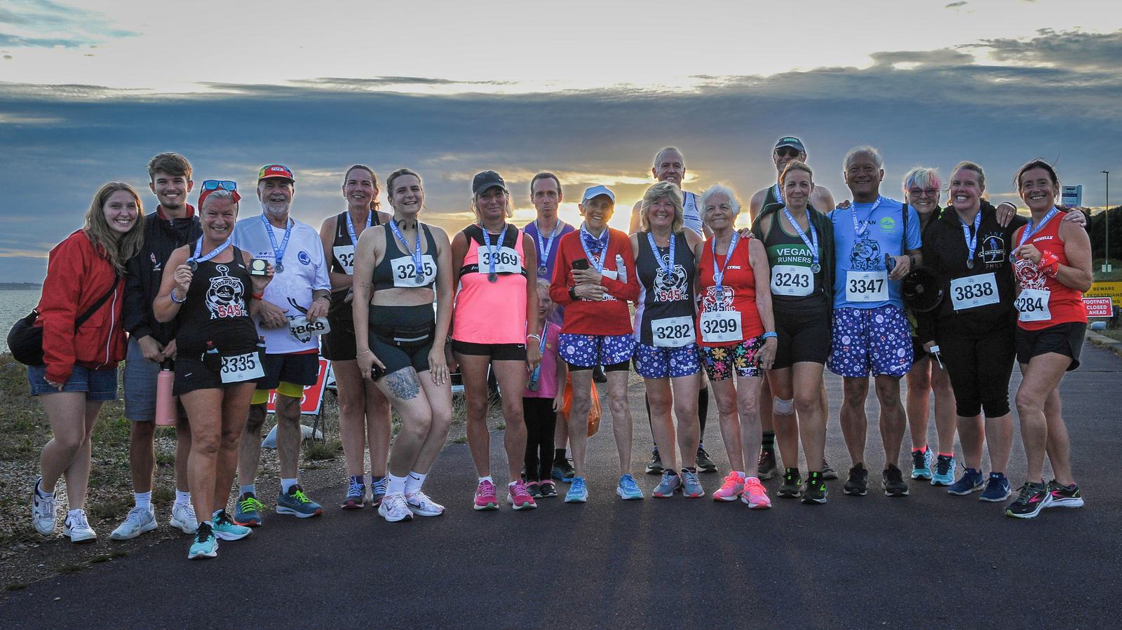A group of runners standing outdoors, each wearing a medal and race number bibs. The background features a cloudy sky with the sun setting, creating a warm glow. The group is dressed in various athletic gear, and most are smiling.
