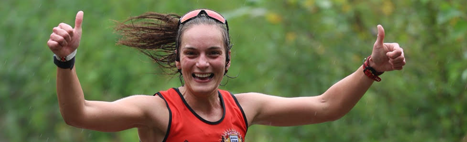 A smiling woman in a red athletic tank top runs outdoors, giving two thumbs up with wet hair and greenery in the background.