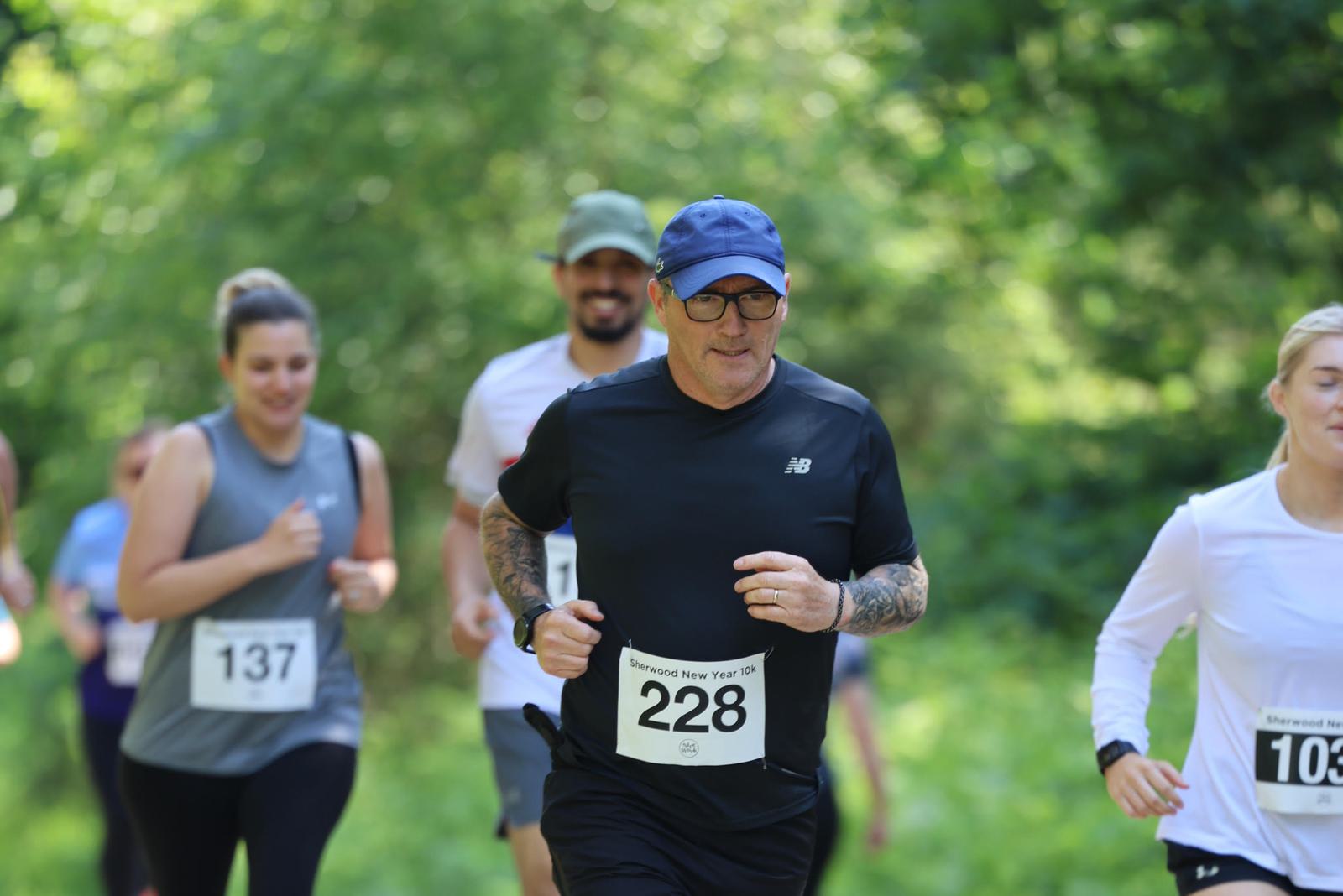 A group of runners in a race, including a man wearing glasses and a cap with bib number 228 in the foreground. They are running on a path surrounded by lush greenery. Other participants are visible in the background.