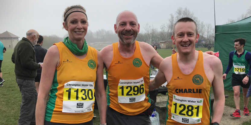 Three runners wearing yellow tops with green logos and race numbers 1130, 1296, and 1281 stand smiling together outdoors. They are participating in the Carsington Water 10K race. A green tent and other participants are visible in the background.