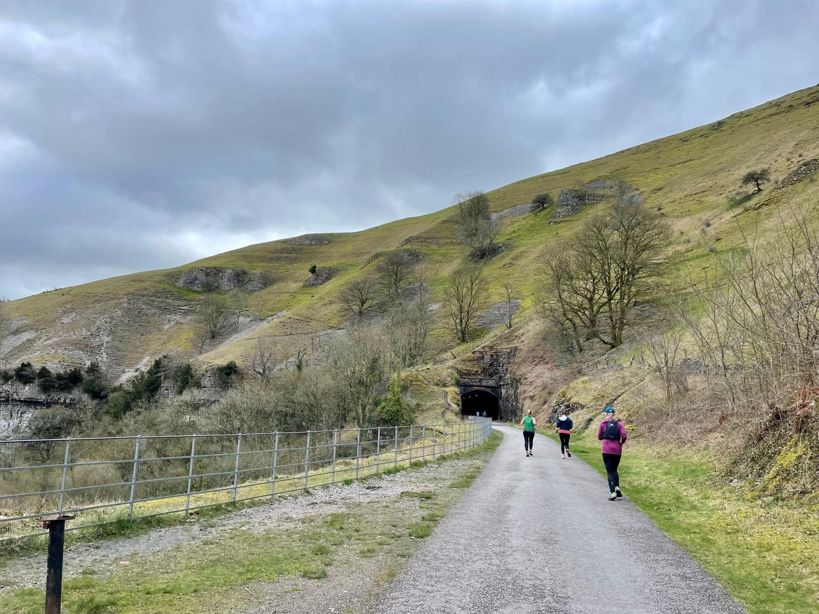 A paved path leads towards a hillside tunnel with three people walking towards it. The surrounding landscape features grassy slopes, scattered trees, and an overcast sky. A metal fence runs alongside the path.