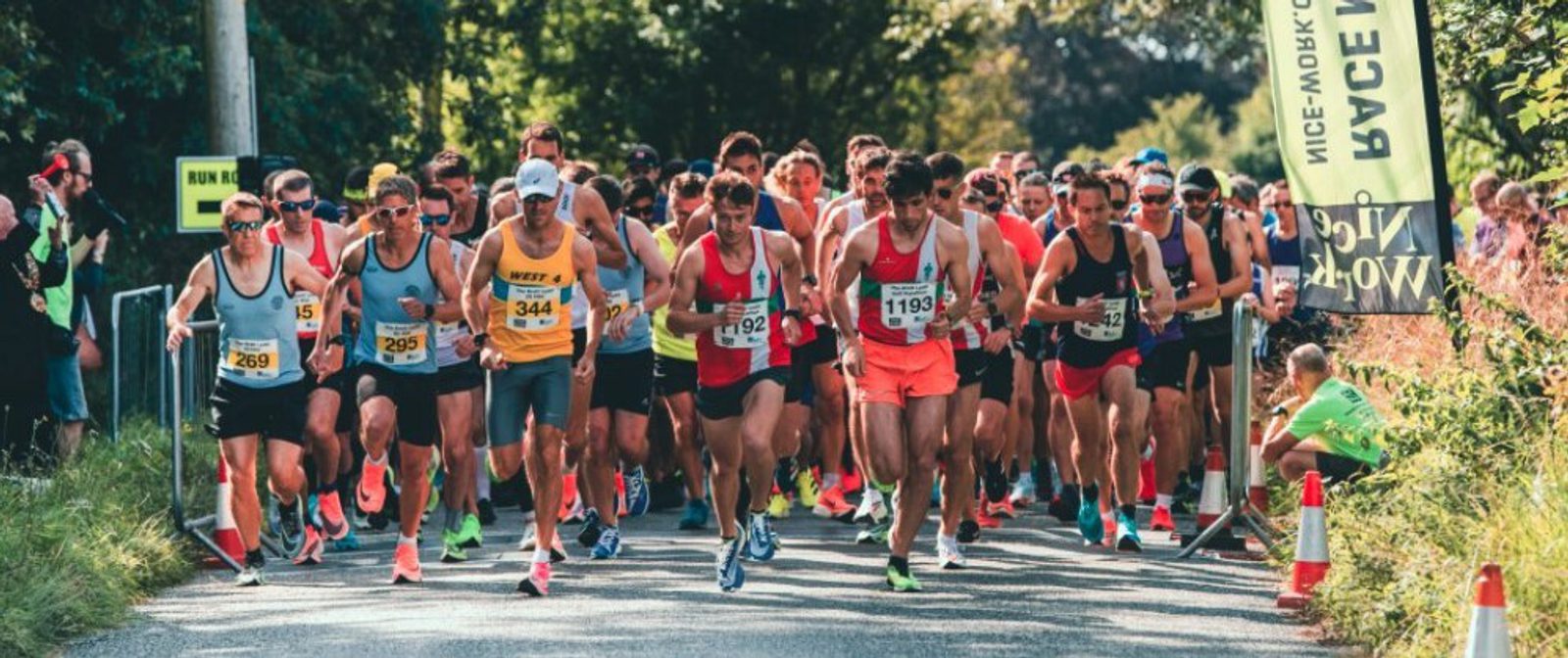A large group of runners, wearing colorful athletic attire and numbered bibs, start a race on a sunny day. Spectators and officials are visible in the background, and a banner and other signage are present along the roadside. Trees and greenery surround the area.