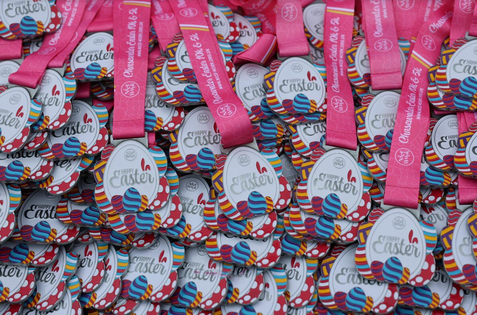 A pile of colorful medals with "Happy Easter" and decorated eggs. Each medal has a pink ribbon labeled "In-person Option A 2.6 & 5k." The medals have vibrant stripes and polka dots in various colors.