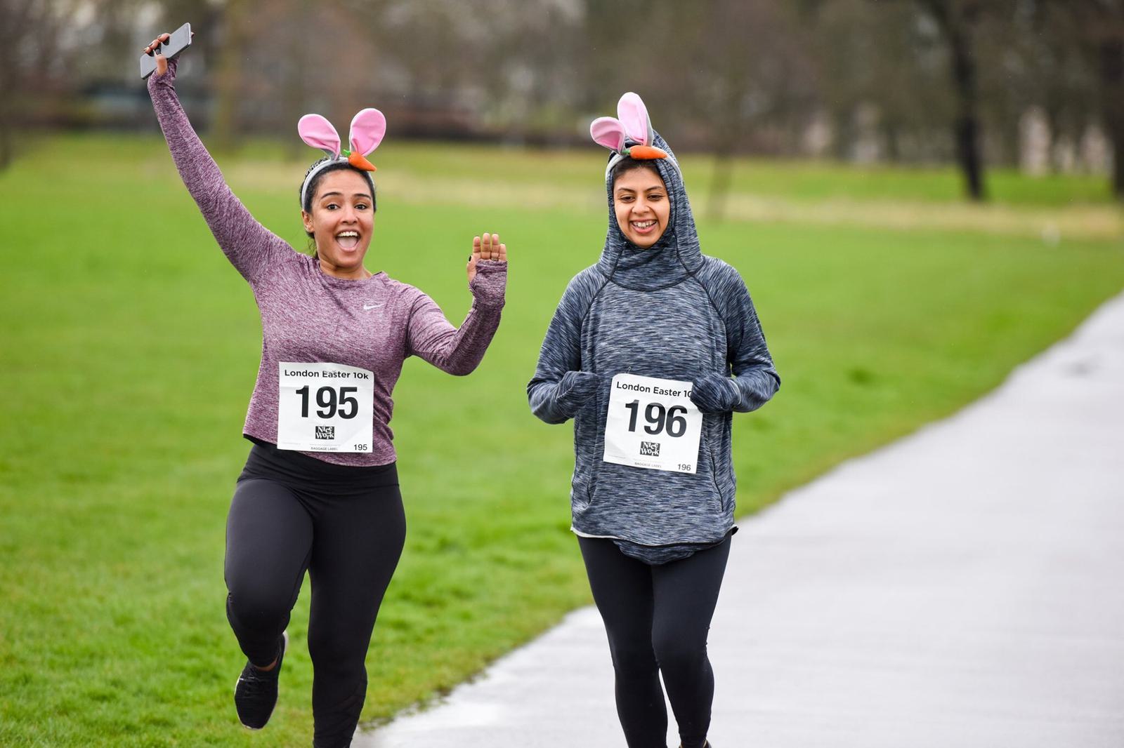Two women wearing bunny ears participate in a race, running on a wet pathway through a park. Both wear numbered race bibs and athletic clothing. One woman is energetically raising a hand, while the other jogs alongside her, smiling.