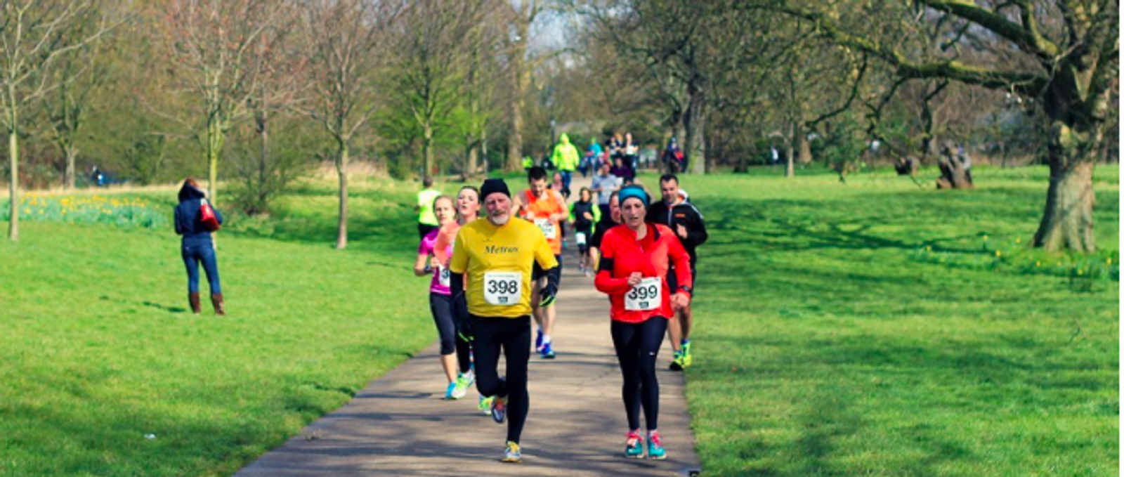 A group of runners, each with a race number, jogs along a park pathway during a race. The greenery of the park surrounds them, with trees on both sides. A few other people are seen walking or running in the background on this sunny day.