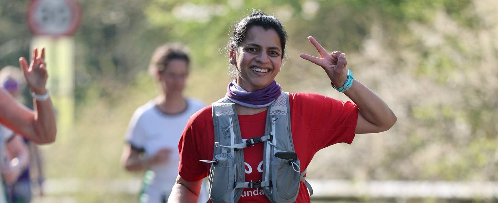 A smiling woman in a red shirt and running gear makes a peace sign with her hand while running outdoors. Other runners and greenery are blurred in the background.