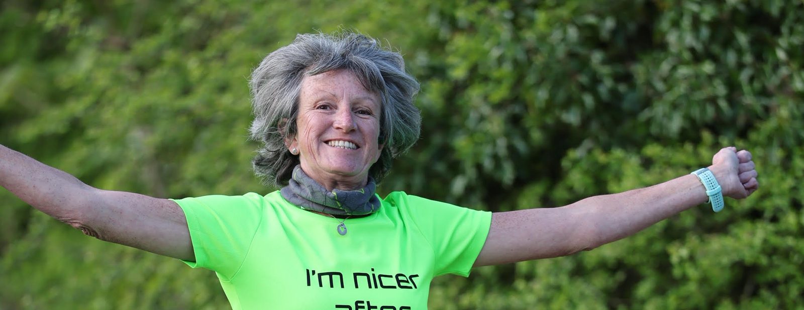 An older woman with gray hair smiles with her arms outstretched, wearing a bright neon green shirt that says "I'm nicer after." Greenery is blurred in the background.