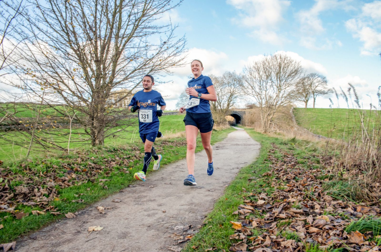 Two runners, wearing race bibs 331 and 326, jog along a leaf-strewn path on a clear day. They are surrounded by grassy fields and bare trees with a tunnel in the background. Both runners are smiling, enjoying the outdoor run.