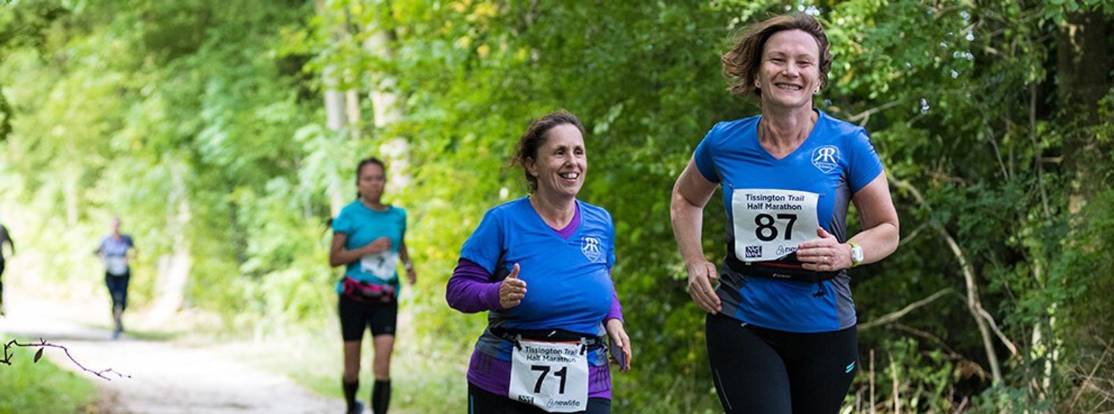 Four women run along a forested trail in a race. Two women in blue shirts, one wearing bib 71 and the other 87, run in the foreground and smile. Two more runners are visible farther back on the path.
