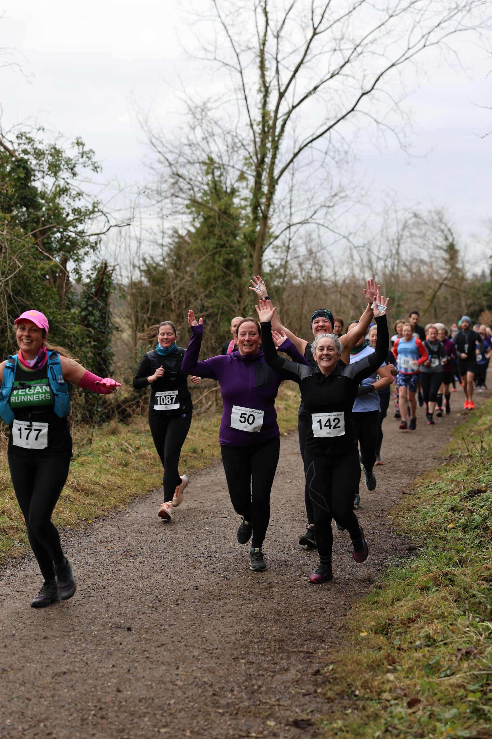 A group of runners on a gravel path during a race. Two women in the center, wearing bibs 50 and 142, are smiling and raising their hands in excitement. Other participants are visible in the background, running through a wooded area on an overcast day.
