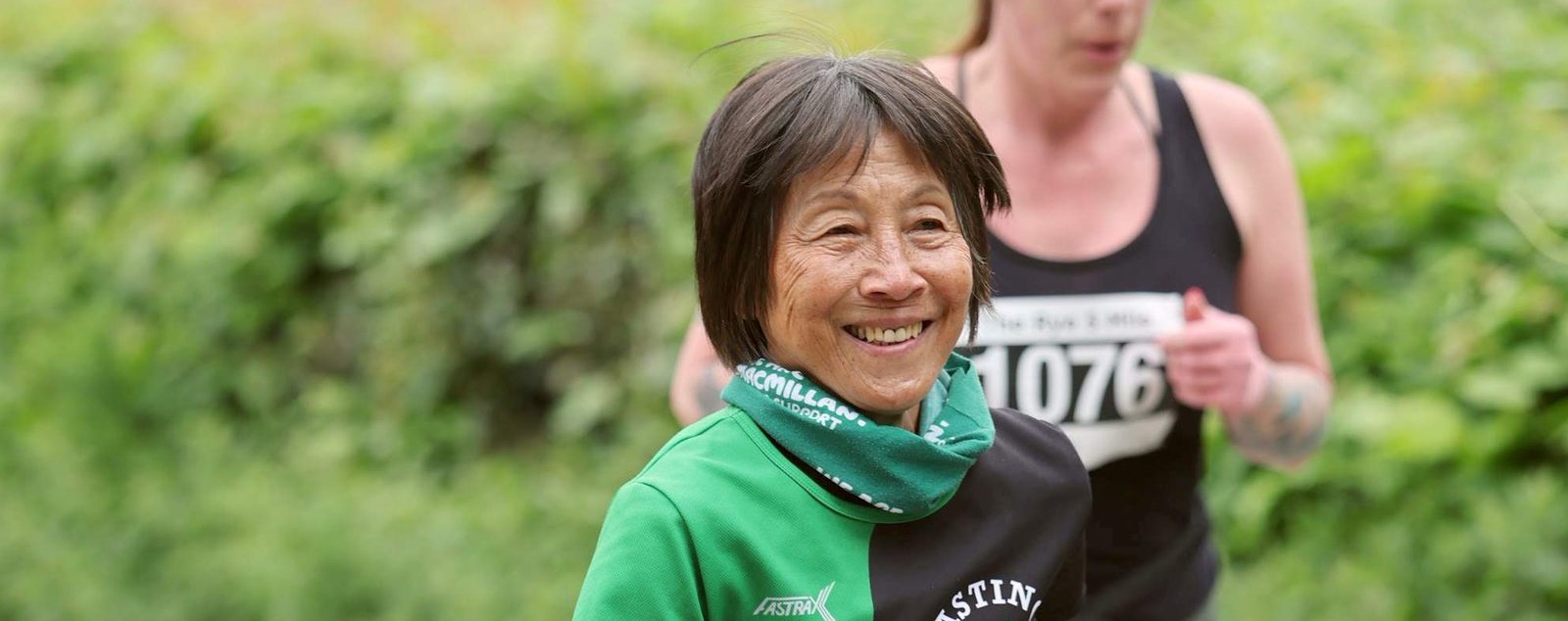 An older woman in a green and black athletic outfit smiles while running outdoors, with another runner slightly blurred in the background. Lush greenery surrounds them.