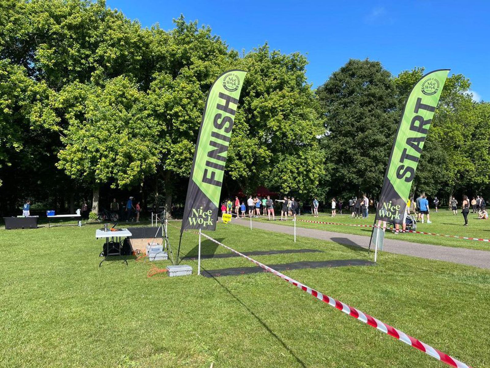 A race event scene in a park with green trees. Two tall flags marked "START" and "FINISH" stand beside each other. Red and white tape cordons off the race area. Participants are gathered in the background near tables under a clear blue sky.