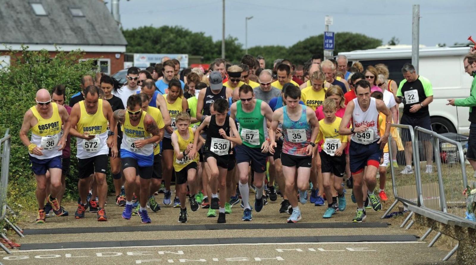 A group of runners, both men and women, are starting a race. They are wearing athletic clothing, many with bib numbers pinned to their shirts. They are at a starting line marked with "GOLDEN MILE." Spectators and a blue sky are visible in the background.