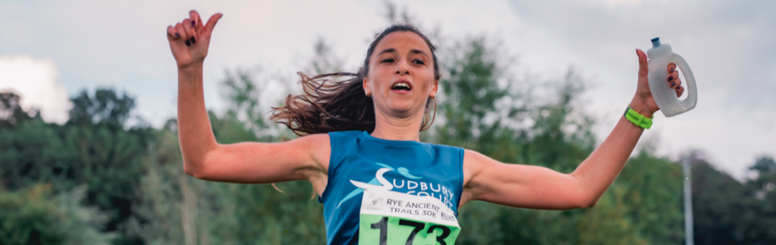 A runner wearing a blue tank top and race bib number 173 celebrates crossing the finish line while holding a water bottle. The runner has long hair flowing behind and appears to be outdoors, with a background of trees and a partly cloudy sky.