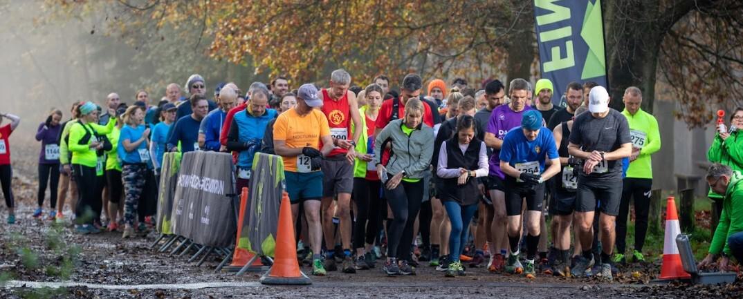 A group of runners at the starting line of a race on an autumn day. Some runners are looking at their watches while others are focused ahead. Trees with fall foliage can be seen in the background, and race banners and orange cones line the path.