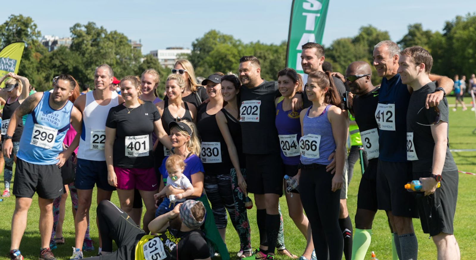 A group of people pose together outside on a sunny day, wearing athletic gear and bib numbers after a race. Some hold water bottles; one person in front has a baby. They are standing on a grassy area with trees and event banners in the background.