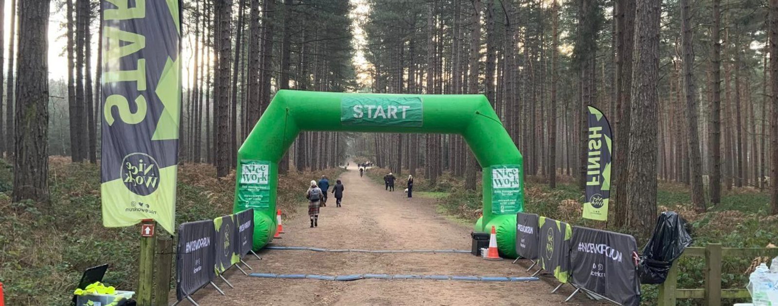 A start line for an outdoor race is set up in a forest. The path is lined with flags and banners, indicating sponsors and directions. An inflatable green arch marked "START" spans the path. Participants are seen in the distance, presumably preparing for the race.