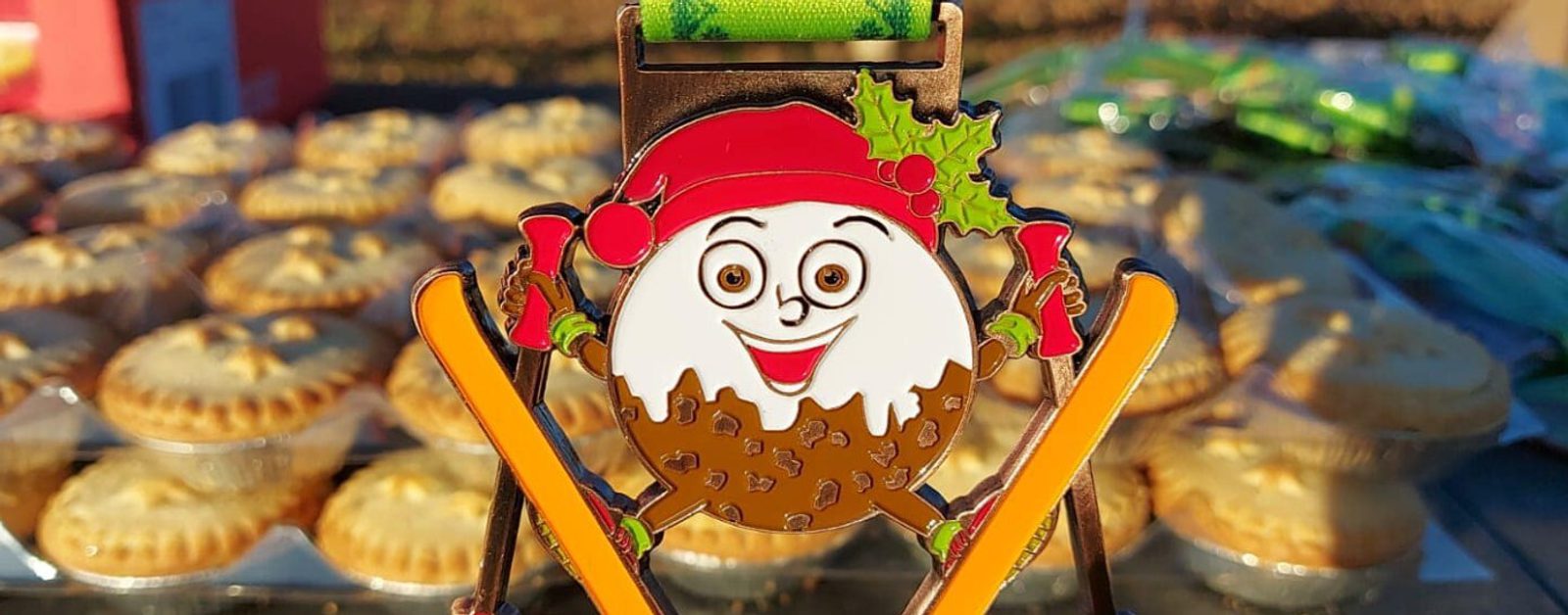 A festive medal featuring a smiling pudding character wearing a red Santa hat and green scarf is held up. The background shows several mince pies on a table, suggesting a holiday-related event or race.