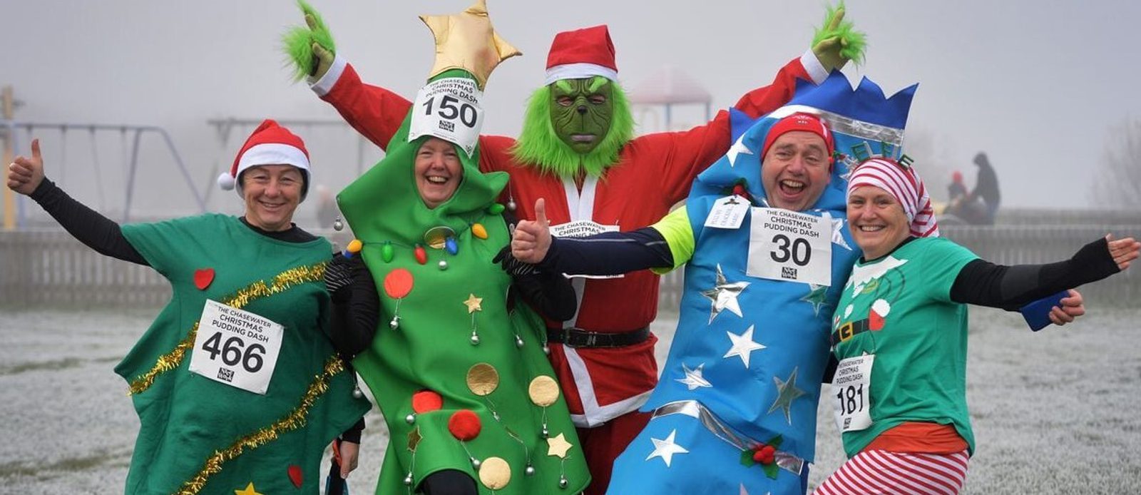 A group of five people dressed in festive costumes, including a Christmas tree, Santa, and an elf, are smiling and posing with their race bibs. The scene is outdoors with a frosty background. Everyone looks joyful and animated.