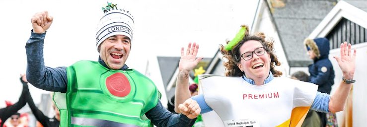 Two people in colorful costumes, smiling and raising their hands while participating in an outdoor event. There are other people and buildings in the blurred background.