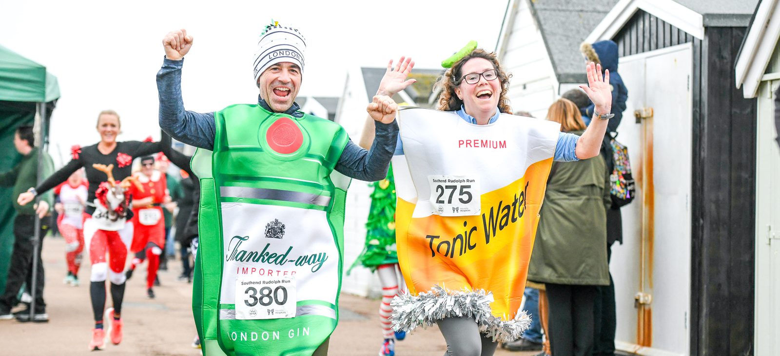 Two people in festive costumes participate in a race; one dressed as a gin bottle and the other as a tonic water can. They are smiling and waving at the camera. Other runners in holiday attire can be seen in the background, and there are sheds and a tent on the side.