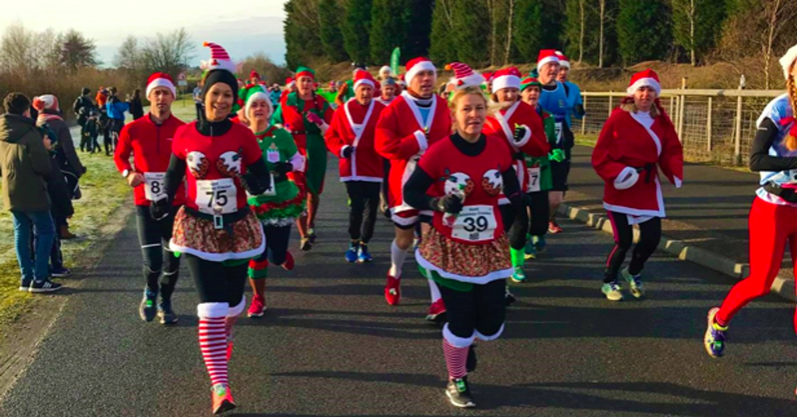 A group of runners dressed in festive holiday attire, including Santa hats and costumes, participate in a race on a paved road. A diverse crowd is seen both running and observing along the sides. Trees and wintry scenery form the background.