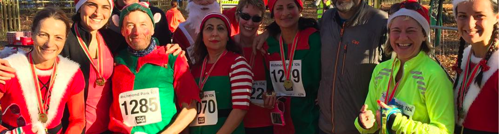 A group of people dressed in festive holiday attire, including Santa and elf costumes, standing together and smiling. They appear to be participants in a race, as some wear numbered bibs and medals. They are outdoors on a sunny day, with trees in the background.