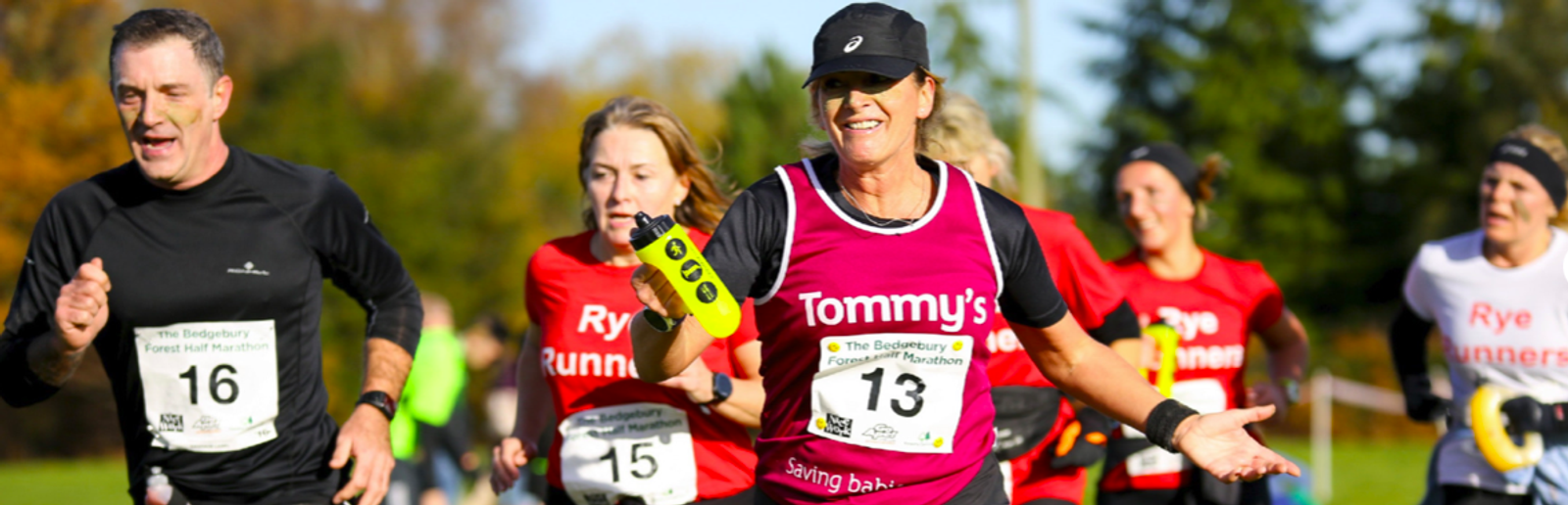 A group of runners in a race, with the runner in the foreground wearing a pink shirt with "Tommy's" written on it and holding a yellow baton. Other runners in red shirts are visible in the background, and they appear to be running through a grassy area.