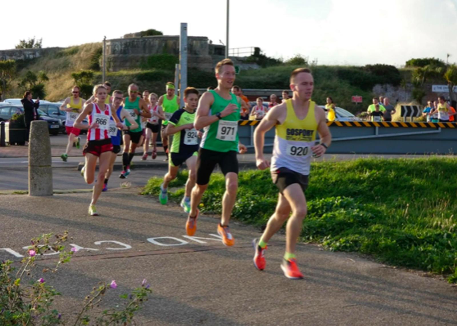 A group of runners competes in an outdoor race. Leading the pack is a runner in a white and yellow tank top with the number 920. Other participants follow closely behind, including runners in numbered bibs ranging from 866 to 990. The race takes place on a clear day.