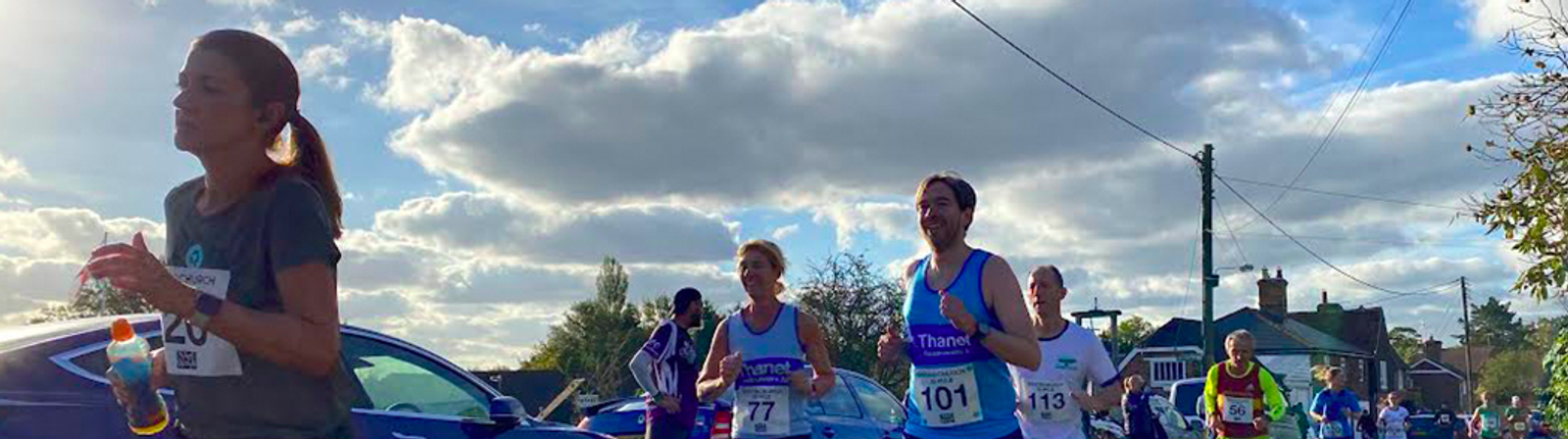 Runners participating in an outdoor race on a sunny day with scattered clouds. They are on a road lined with parked cars and residential houses. The runners appear focused, with some wearing numbered bibs. Trees and utility poles are also visible in the background.