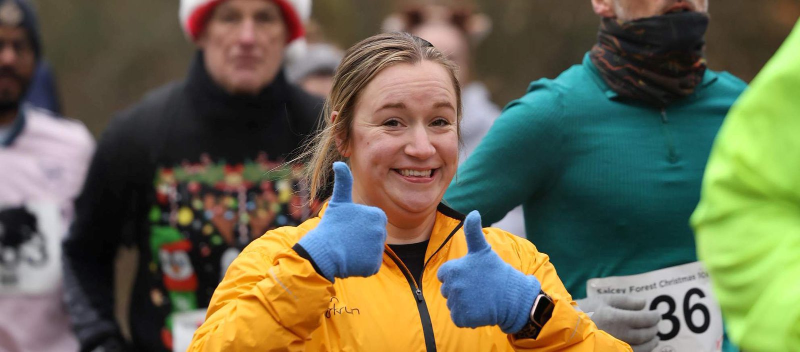 A smiling woman in a yellow jacket and blue gloves gives two thumbs up while running in a race, surrounded by other participants wearing race numbers and winter clothing.