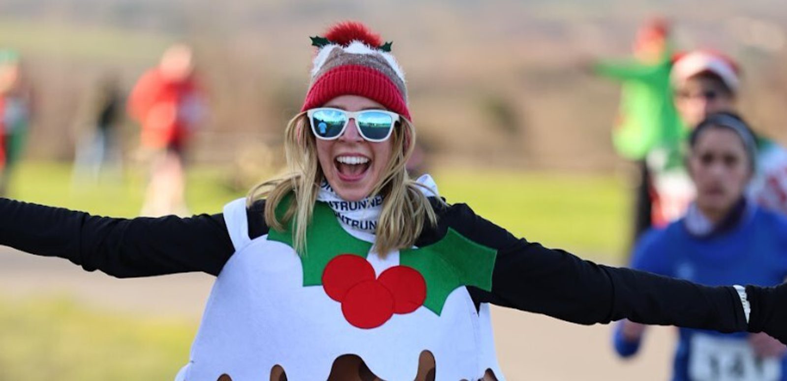 A woman wearing sunglasses, a festive hat, and a Christmas pudding costume smiles with arms outstretched while participating in an outdoor race. Other runners are blurred in the background.