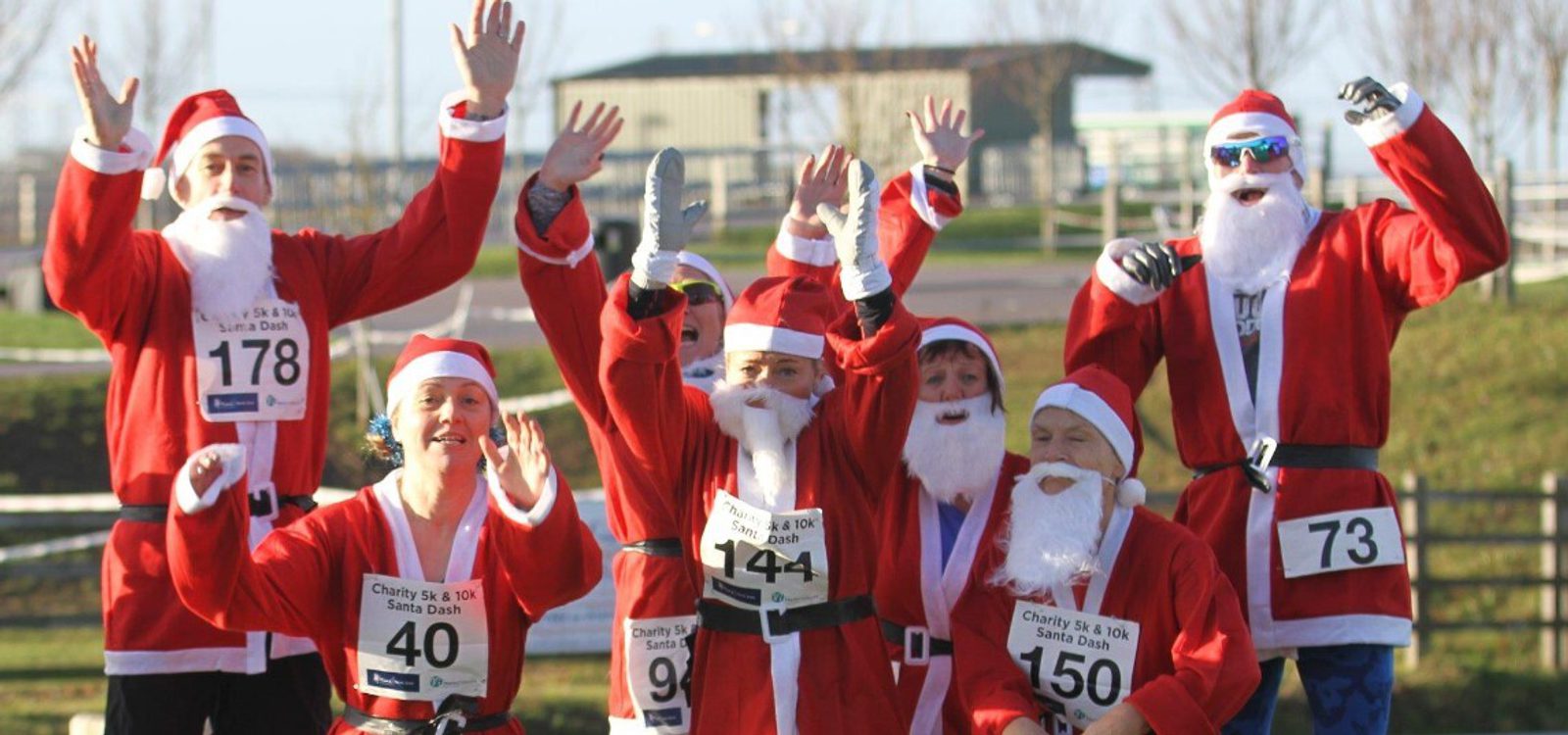 A group of people dressed as Santa Claus, wearing red suits and white beards, cheerfully wave their hands in the air. They have race bibs with numbers on their chests, suggesting they are participating in a charity run. The background features an outdoor setting.