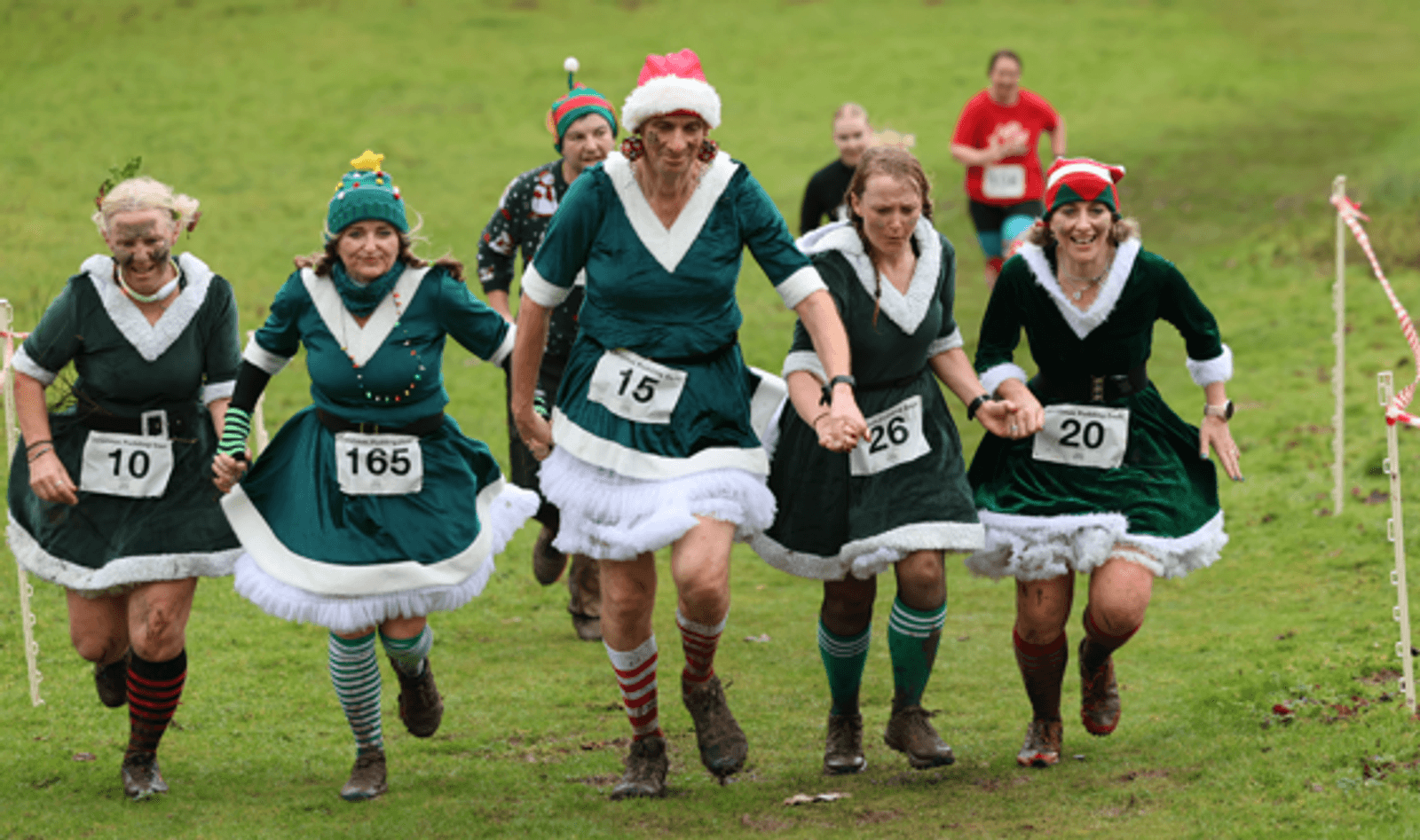 Five people in green elf costumes, striped socks, and Christmas hats are running through a grassy field in a race, holding hands and smiling. Other participants are visible in the background. Race numbers are pinned to their outfits.