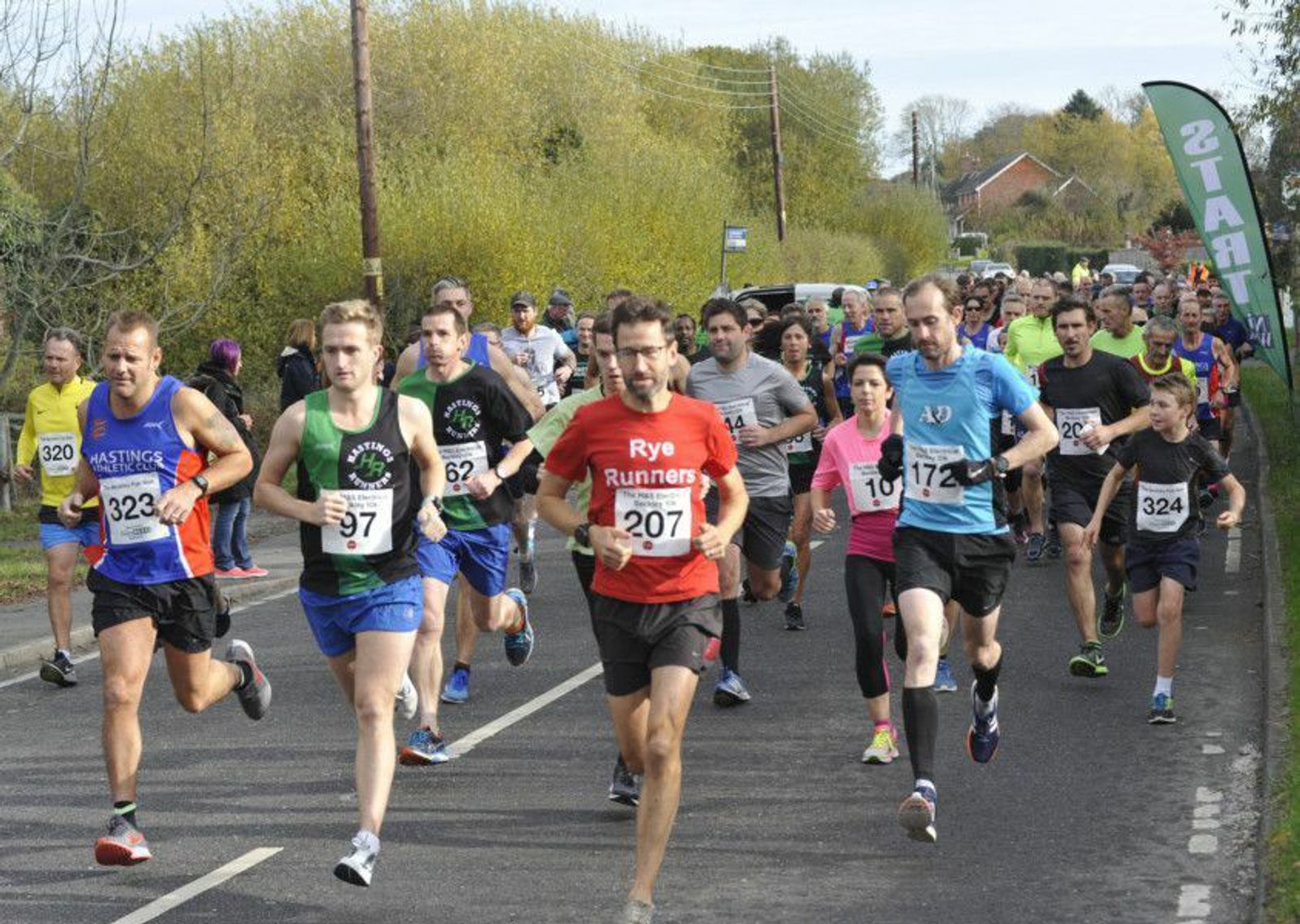 A large group of runners, wearing race numbers on their shirts, participate in a road race on a two-lane road. Trees and houses are visible in the background. Many participants are dressed in colorful athletic gear, and a sign reading "START" is visible on the right.